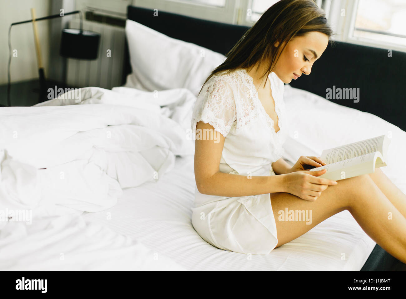 Young pretty woman sitting on a bed and reading a book Stock Photo - Alamy