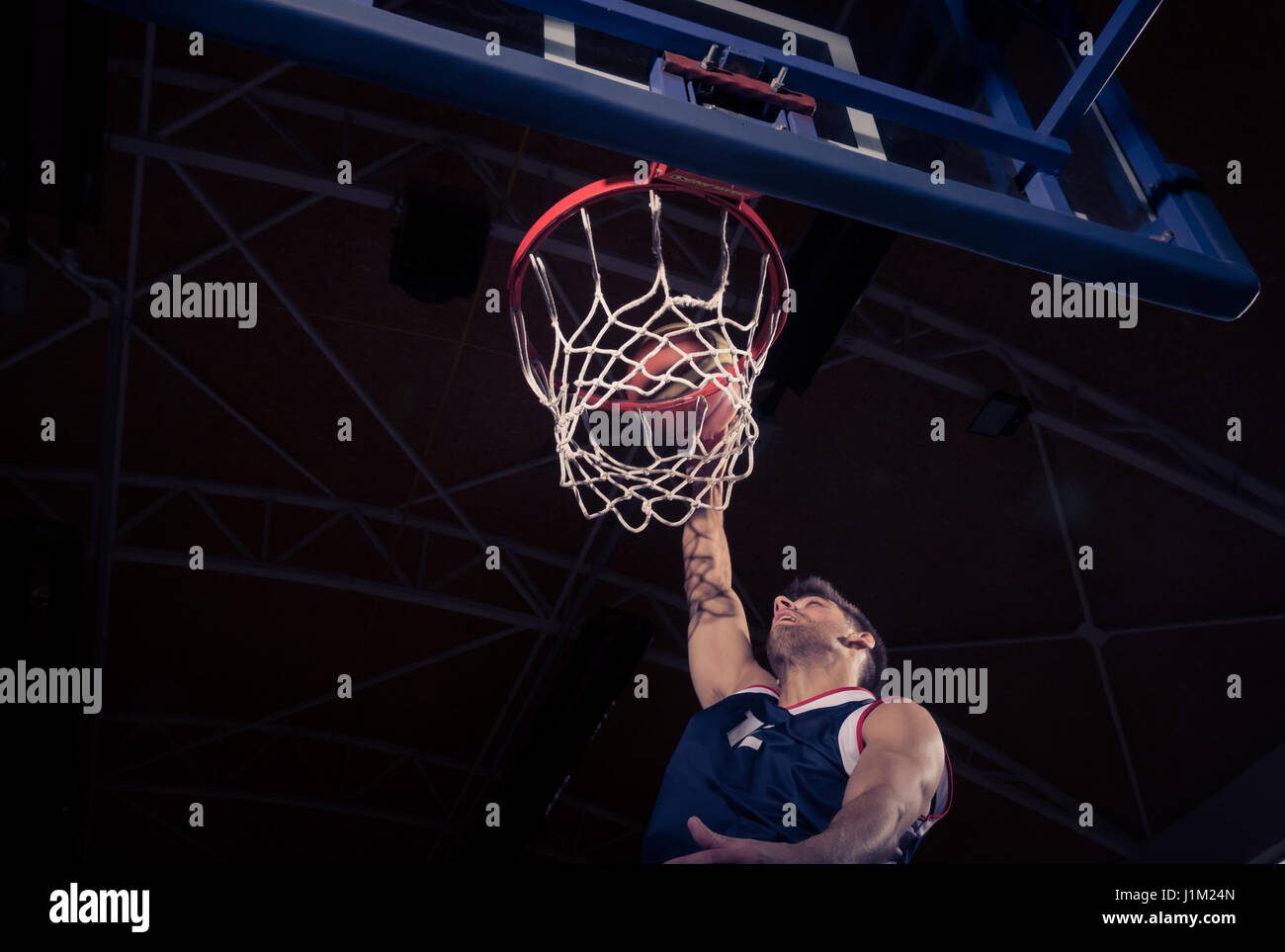 one young adult man, basketball player, one hand, net rim, low angle view, slam dunk, dark indoors. Stock Photo