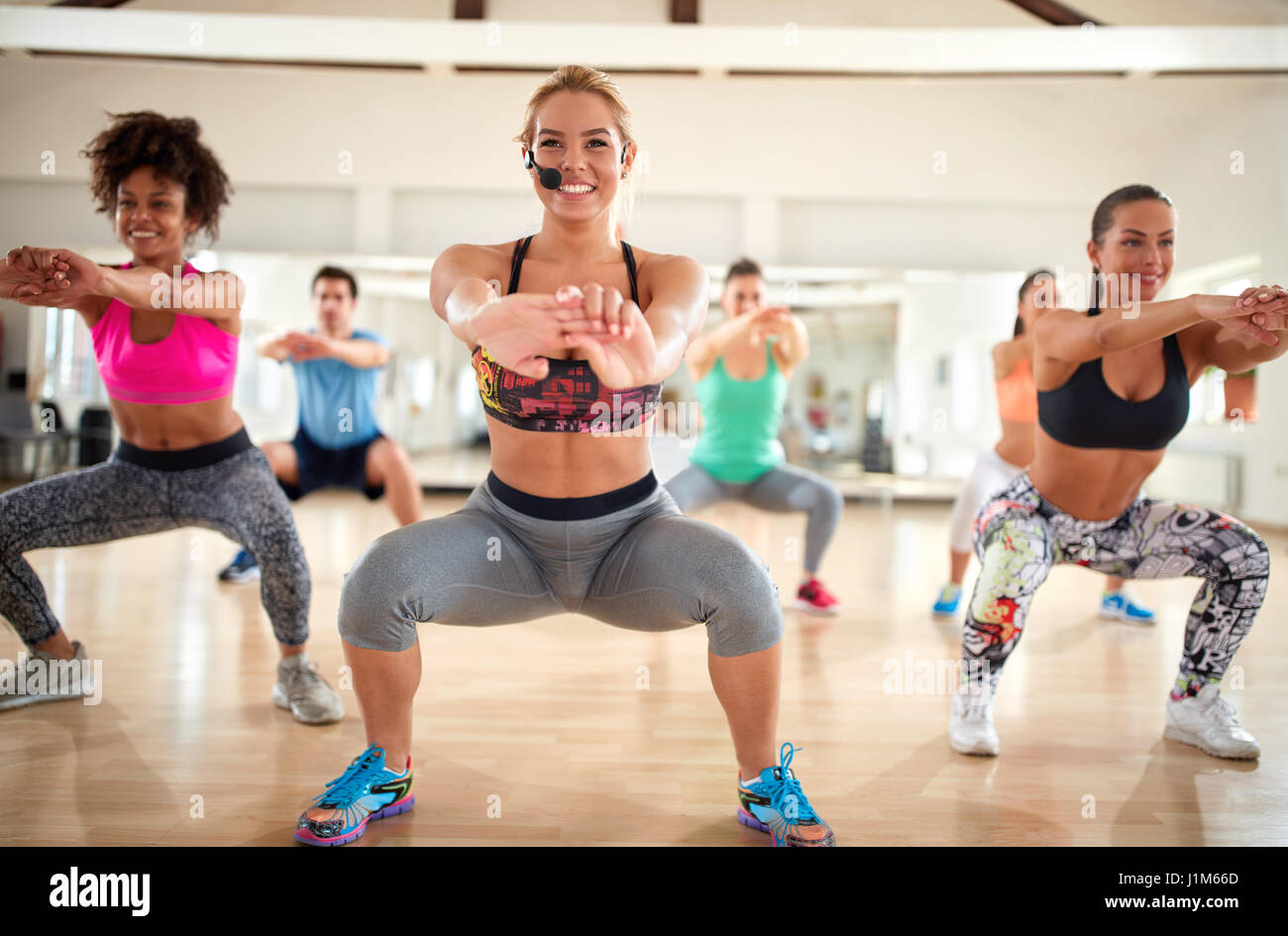 Smiling blond female trainer with headset doing squats with fitness group indoor Stock Photo