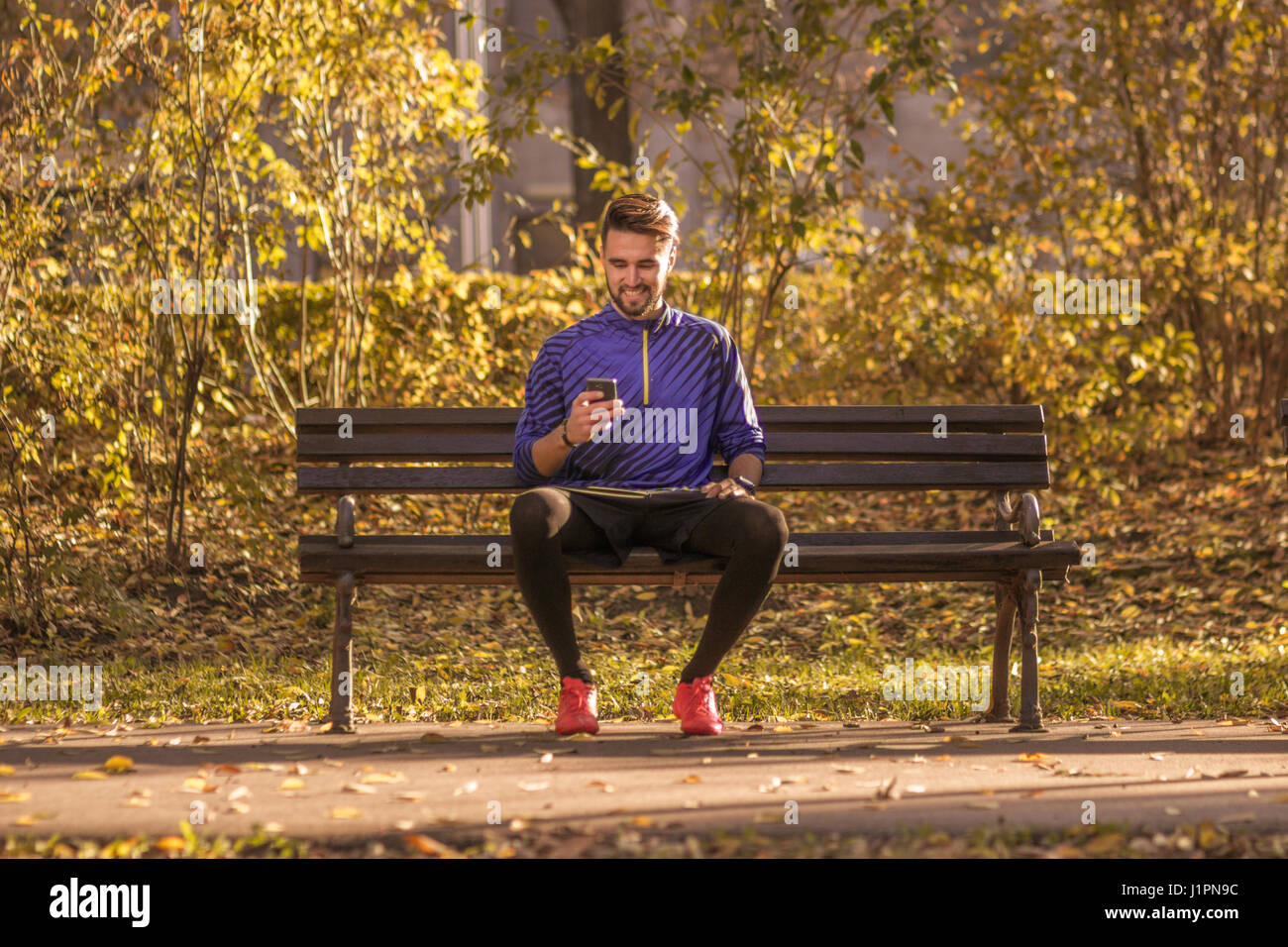 young man sitting bench park athlete, football player, soccer player, using smartphone. autumn leaves orange, yellow colors. Stock Photo