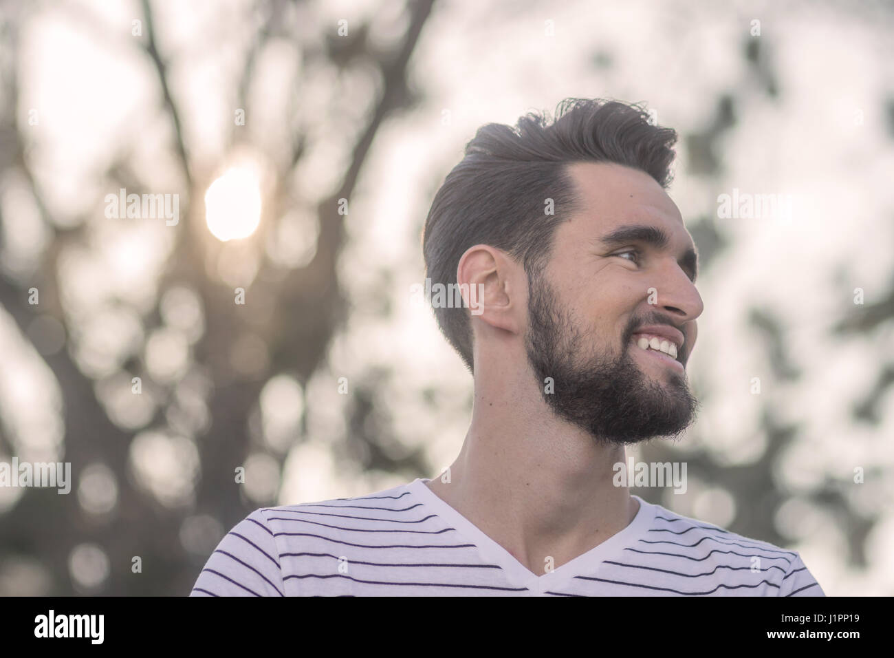 One smiling young adult man, looking sideways, head, face close up. Wearing t-shirt, outdoors nature. Low angle shot. Sun. Stock Photo