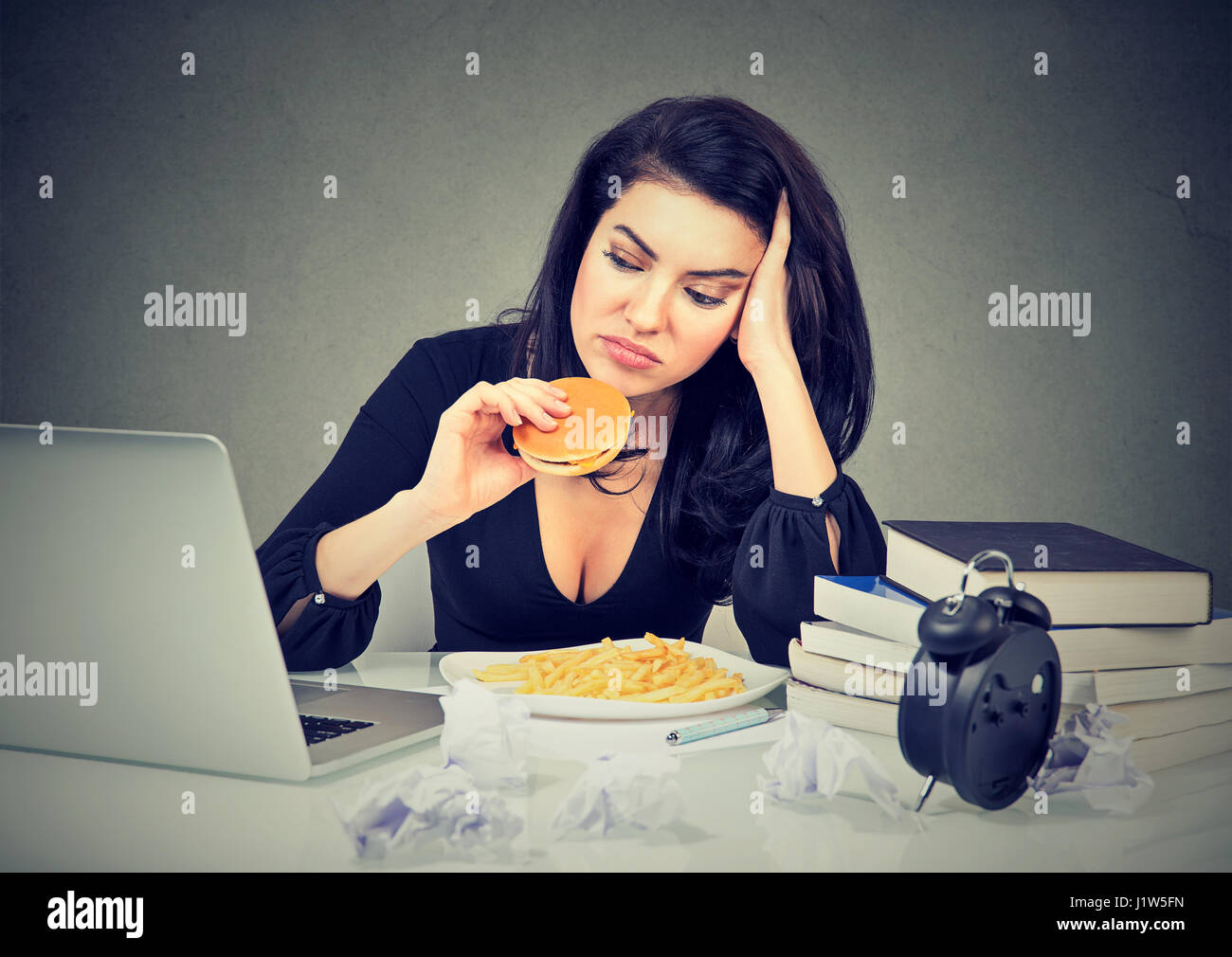 Sedentary lifestyle and junk food concept. Tired stressed woman sitting at her desk eating hamburger and french fries Stock Photo