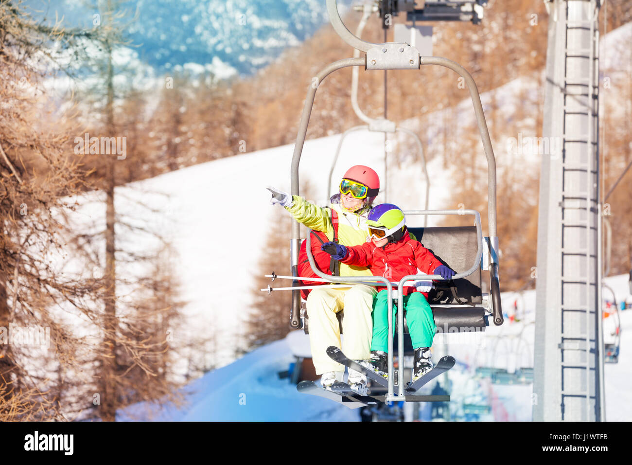 Active woman and her son lifting on chairlift,  pointing to snowy peaks with their fingers Stock Photo