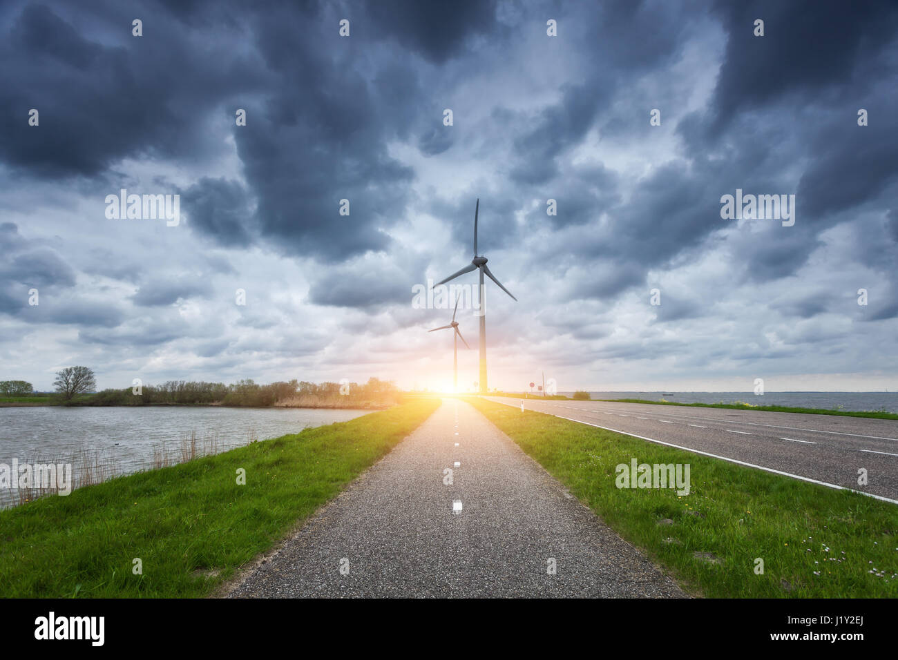 Beautiful asphalt road with wind turbines generating electricity at sunset. Windmills for electric power production. Landscape with road, green grass  Stock Photo
