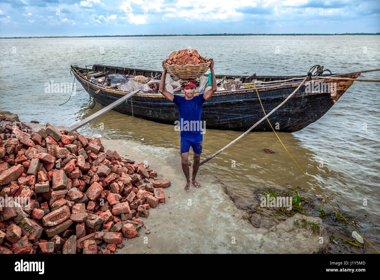 A worker transporting pieces of red brick from a wooden boat onto the Rupnarayan riverbank for river erosion control project. Stock Photo