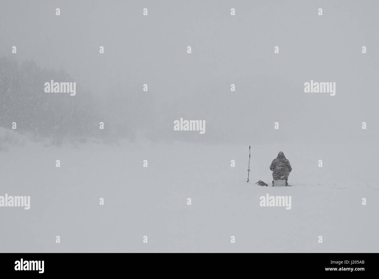 A view of the lone fisherman sitting on the icebound river and fishing during a heavy snowfall Stock Photo
