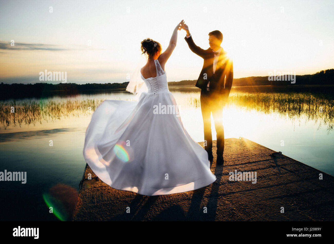 Bride is spinning in a white dress holding hand the groom on the bank of the lake at sunset with sunshine and sunlight Stock Photo