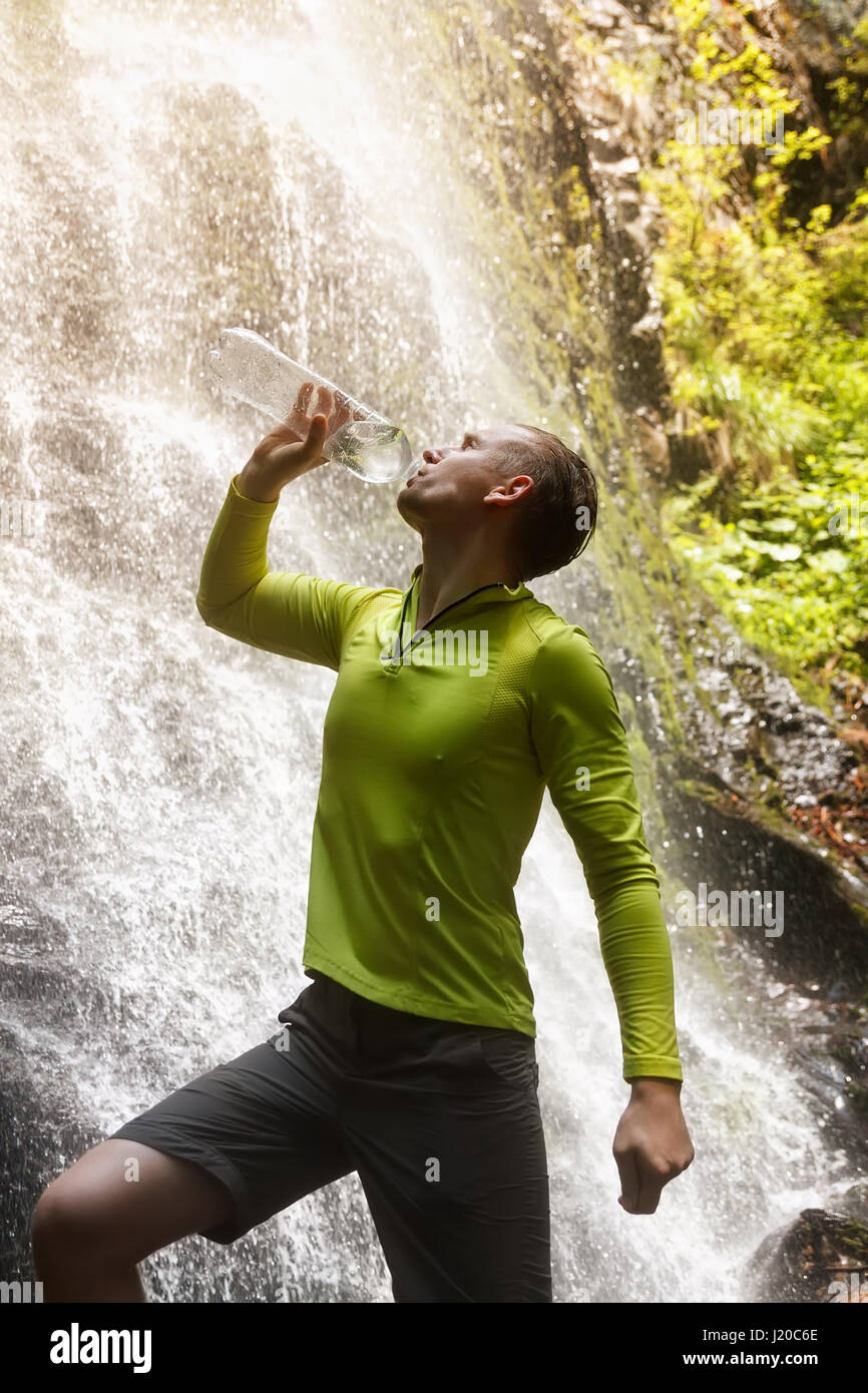 hiker man drinking fresh clean water from a natural. Young tourist drinking water from a bottle outdoor waterfall in the background. Drink water, hydr Stock Photo
