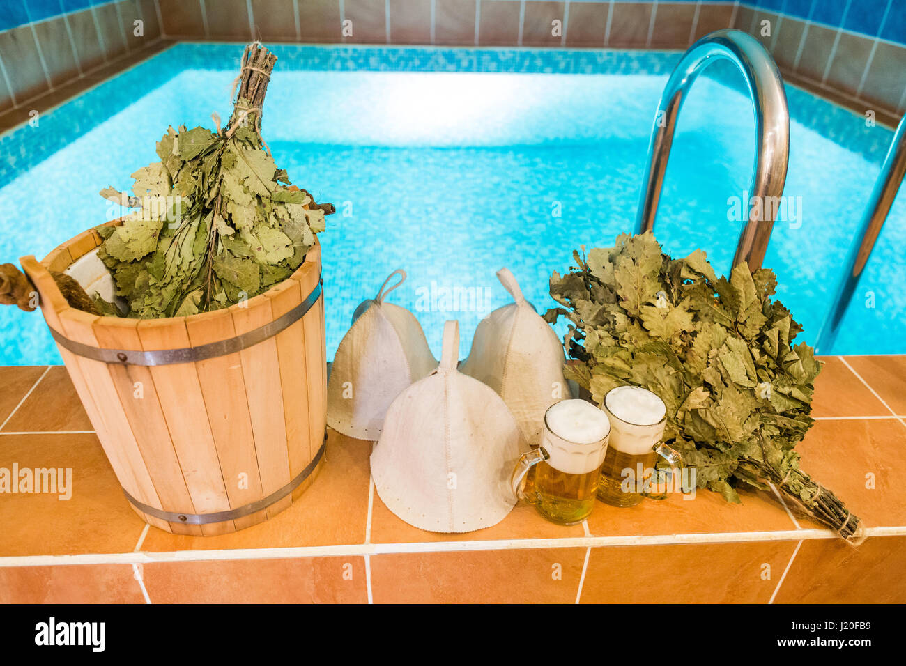Bath accessories in the Russian bath. Bathroom items of traditional Russian sauna. two mugs of light beer, bathing caps, bath brooms from oak leaves against the background of the pool. Stock Photo