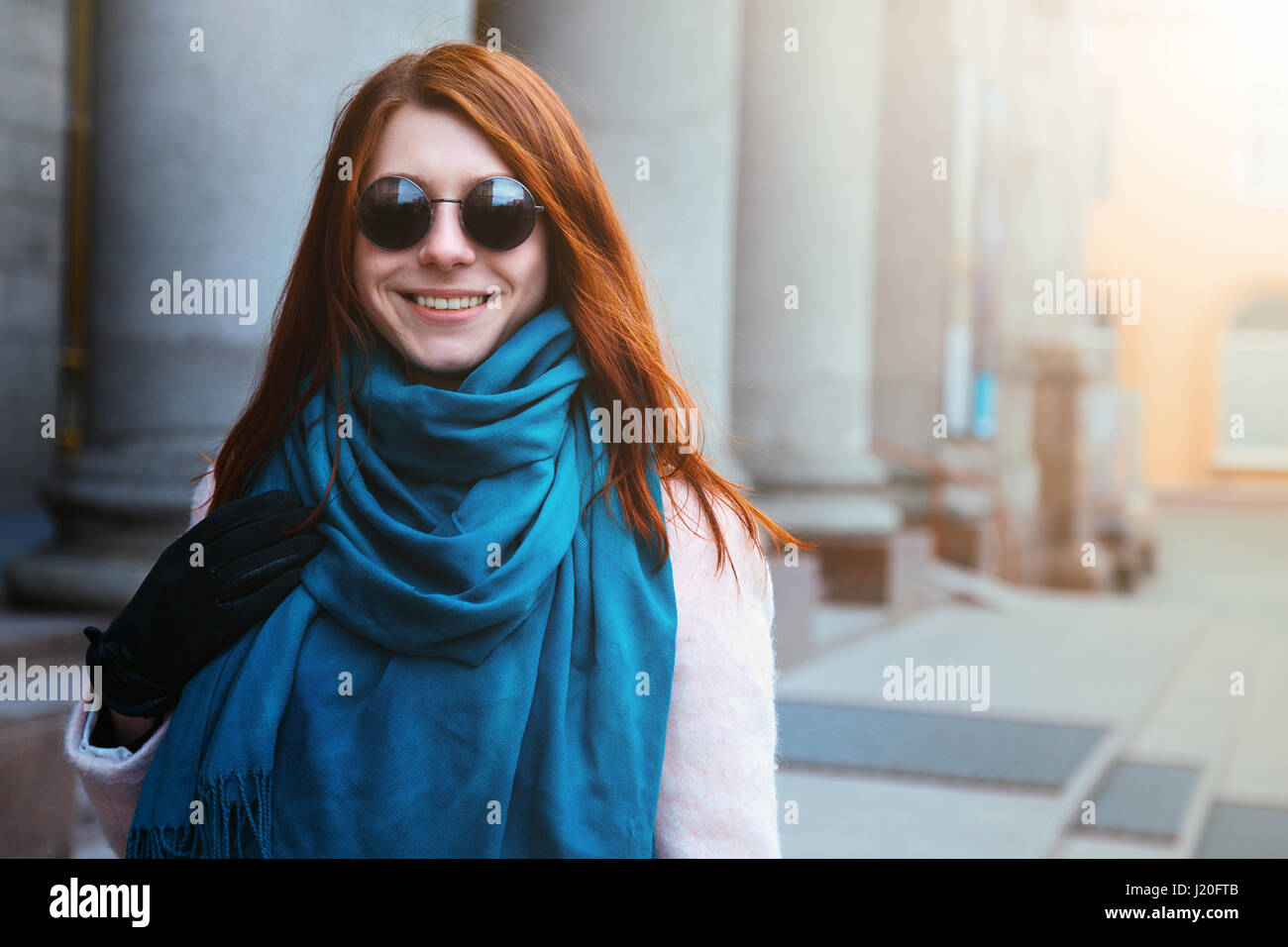 Red haired beautiful girl is walking in the urban background in a pink coat and blue scarf, with sunglasses. Stock Photo