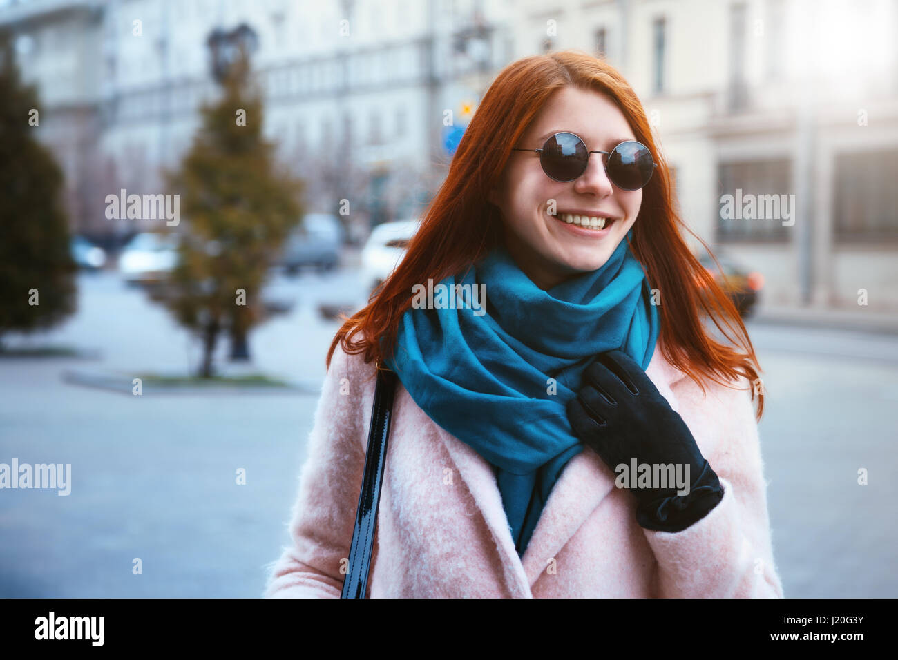 Red haired beautiful girl is walking in the urban background in a pink coat and blue scarf, with sunglasses. Stock Photo