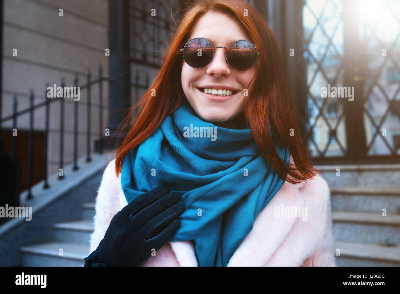 Red haired beautiful girl is walking in the urban background in a pink coat and blue scarf, with sunglasses. Stock Photo