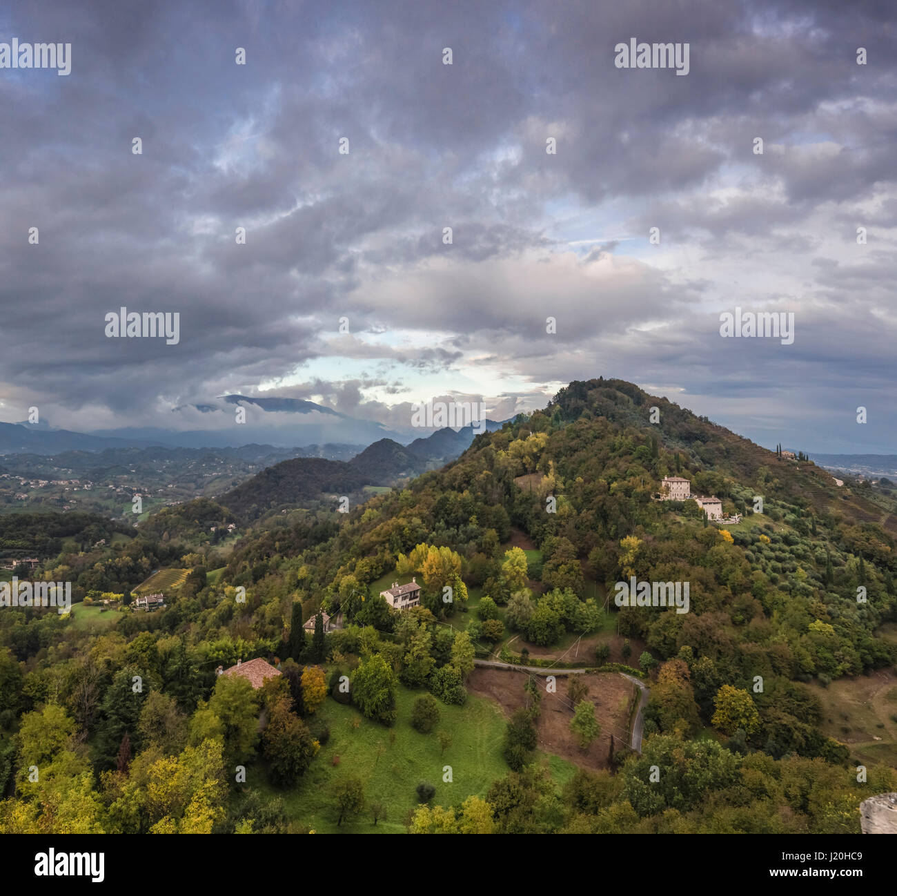 A hilltop in rural Veneto, Italy. Small farmhouses and forest surround with a dramatic sky and mountains in the background. Stock Photo