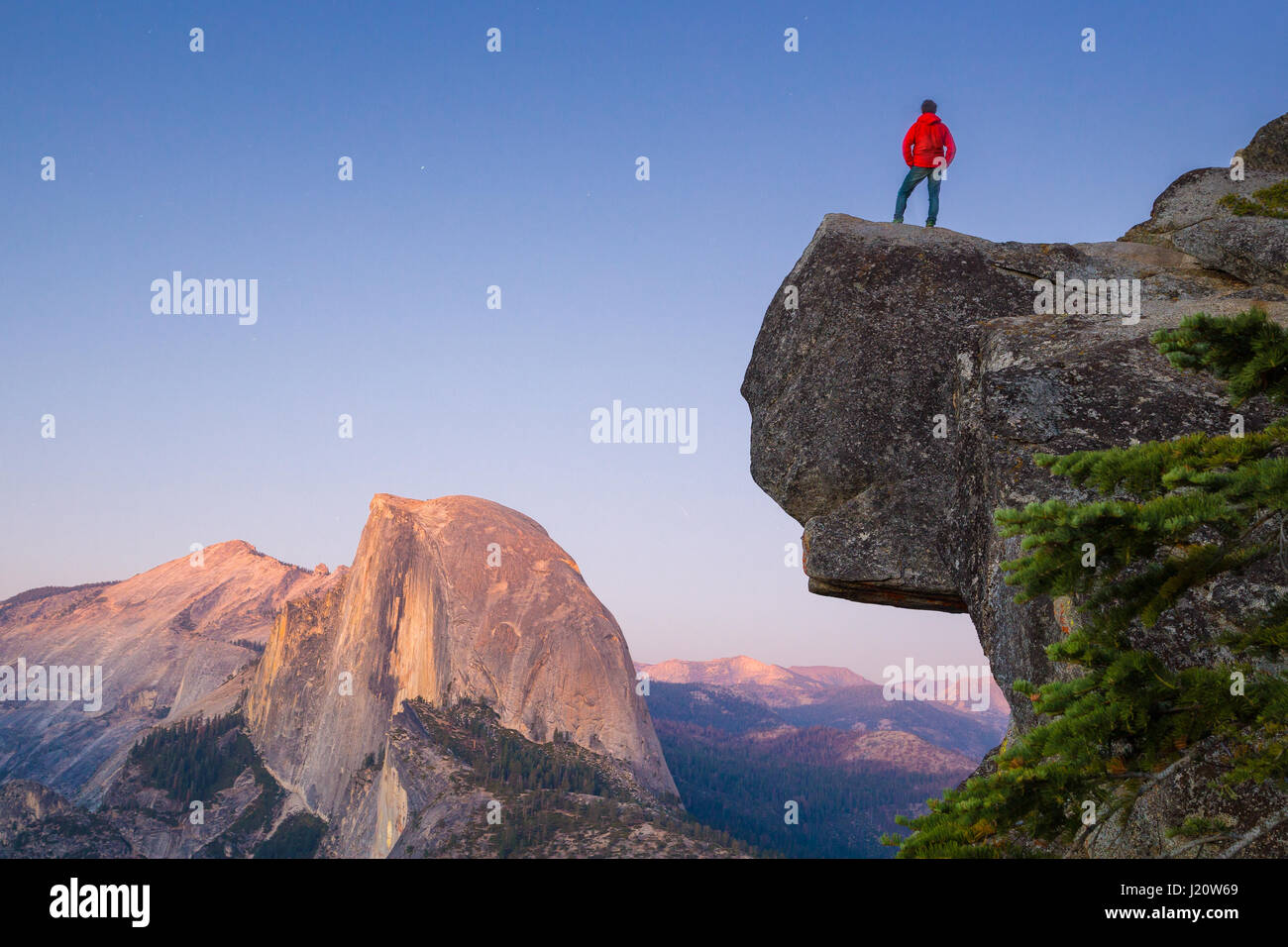 A fearless hiker is standing on an overhanging rock enjoying the view towards famous Half Dome at Glacier Point overlook at sunset, Yosemite NP, USA Stock Photo