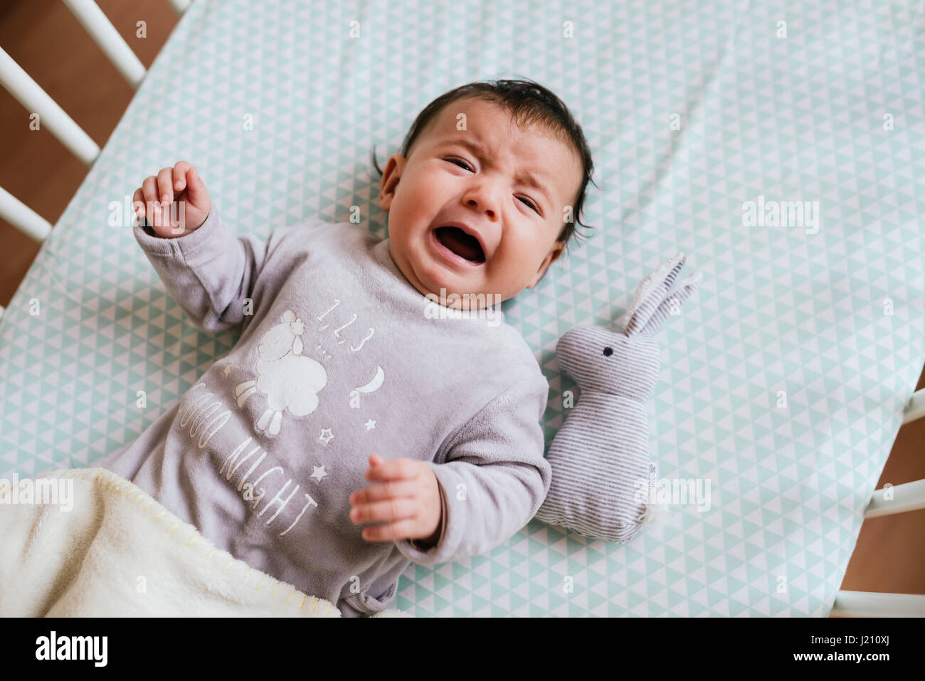 Portrait of crying baby girl lying in crib with toy bunny Stock Photo