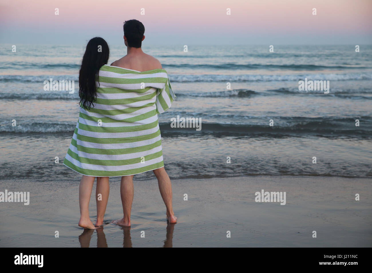 Back view of young couple on the beach wrapped together in towel watching sunset Stock Photo