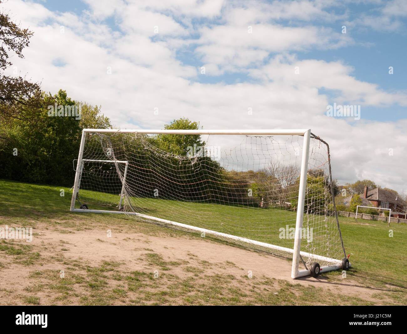 An Empty and Unused Goal Post with A White Net in the Middle of A Park with Grass and Soil on the Ground, Wheels on the Frame to Help Move it and Hous Stock Photo