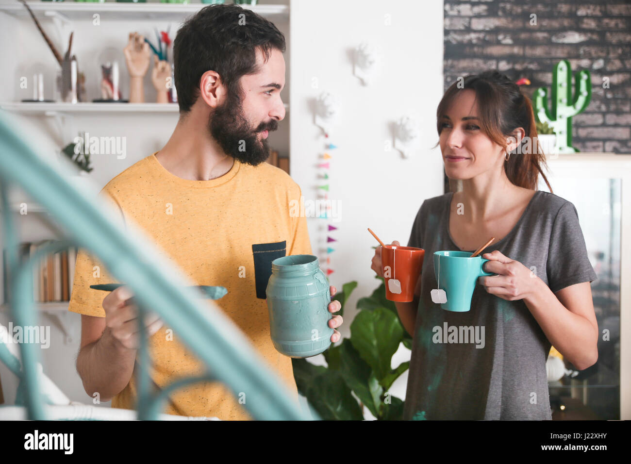 Young woman bringing two cups of tea while her boyfriend painting an armchair Stock Photo