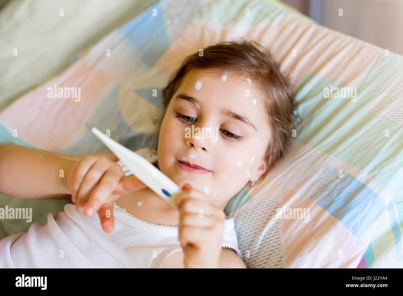 Girl having chickenpox lying in bed looking at clinical thermometer Stock Photo