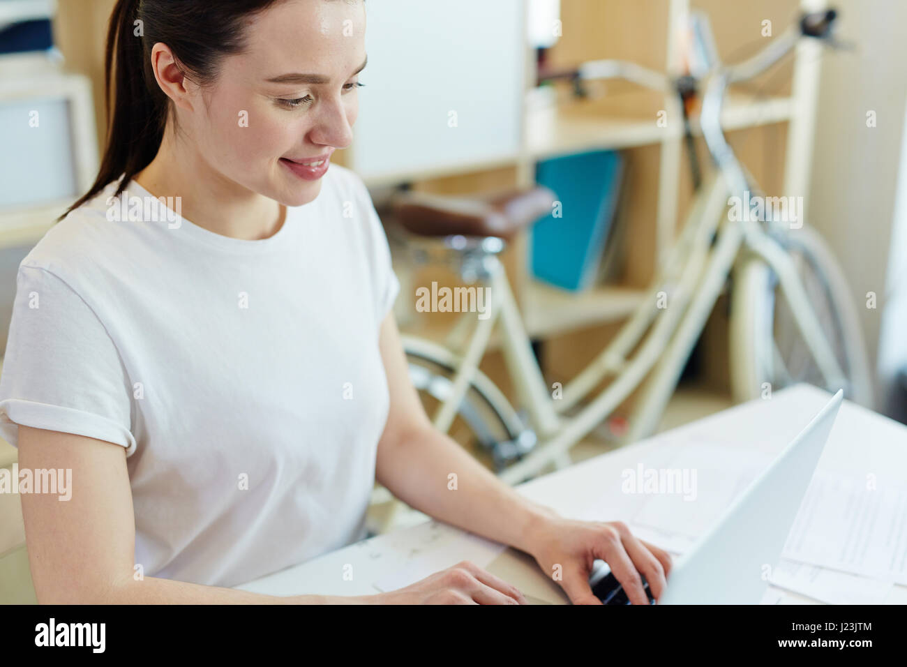 Portrait of pretty young woman dressed in casual clothes working with laptop and smiling in creative office space Stock Photo