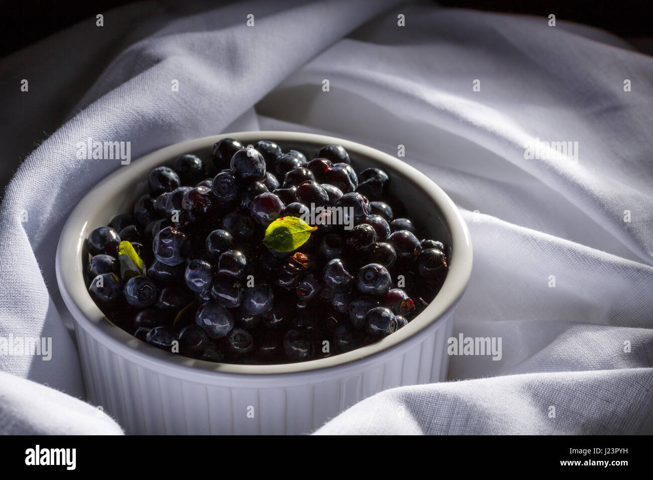 Pile blueberries in white ceramic bowl stay on white organic material. Stock Photo