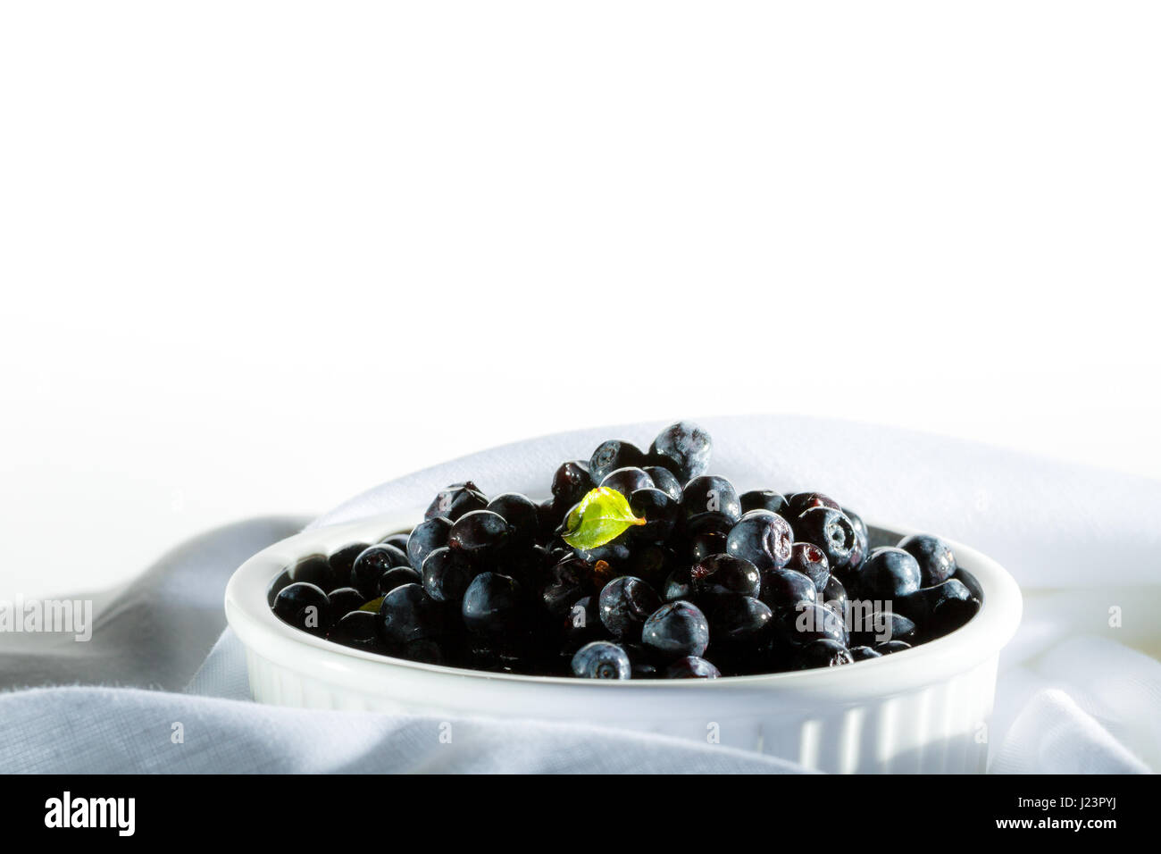 Pile blueberries in white ceramic bowl stay on white organic material. Stock Photo