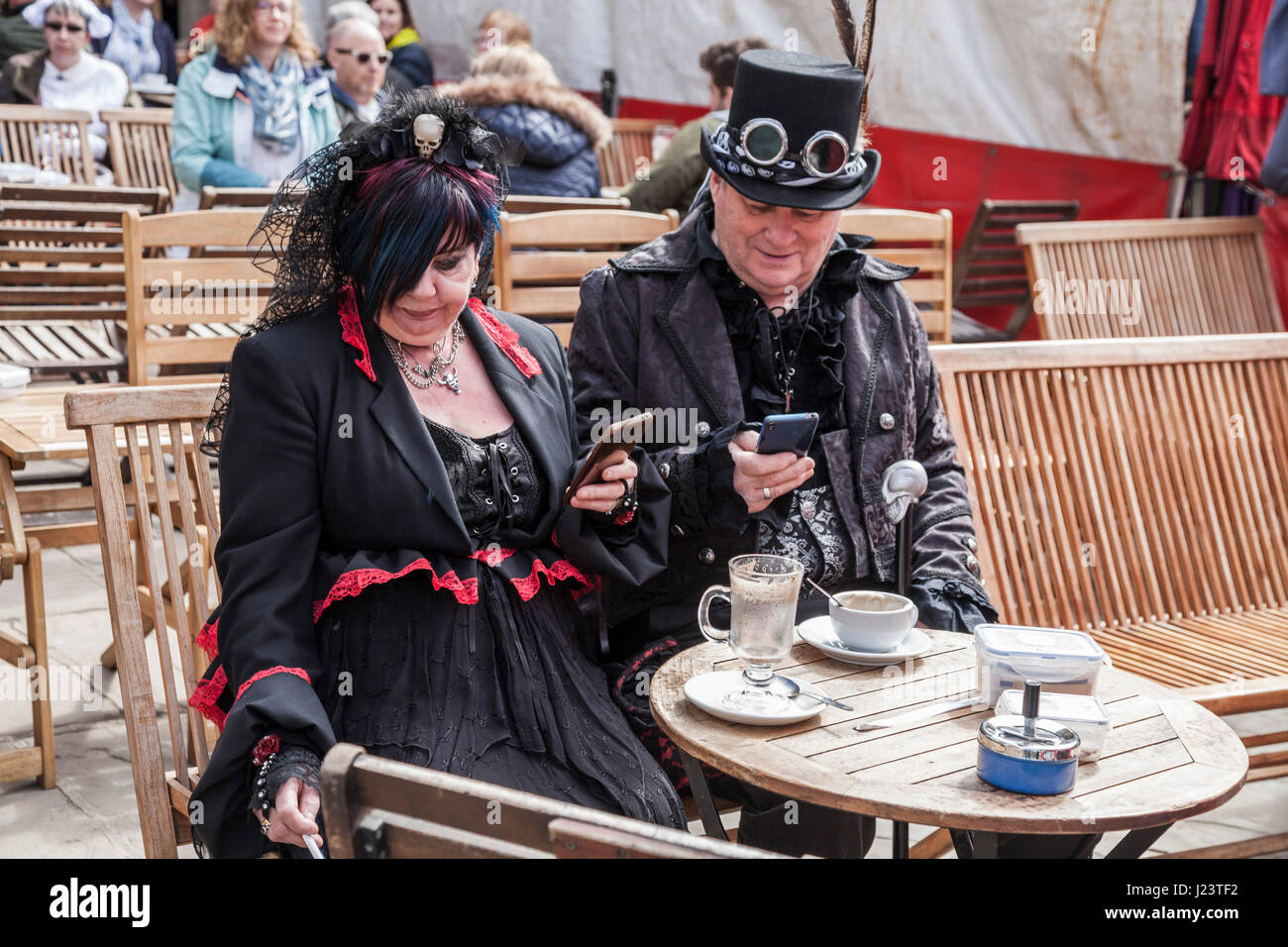 A man and woman dressed up for the Whitby Goth celebrations take a break and sit down for a drink and smoke and look at their phones Stock Photo