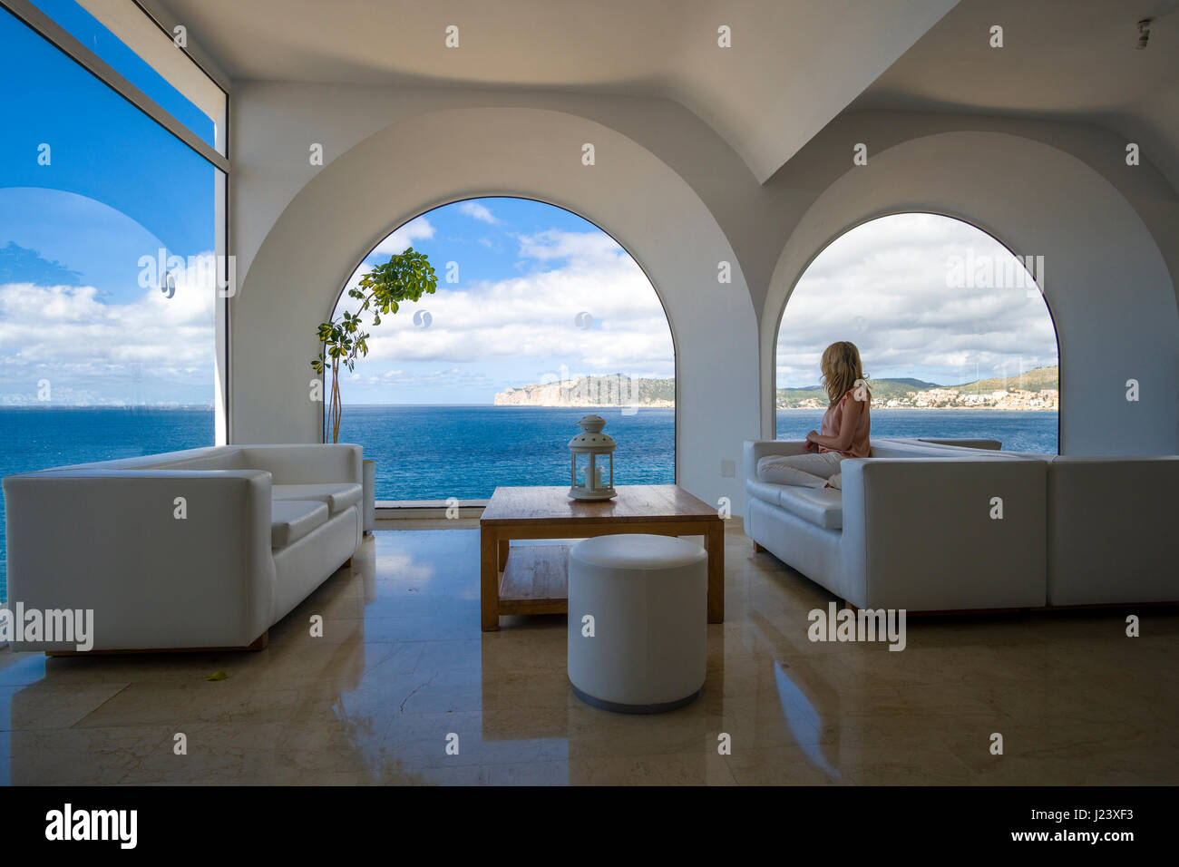 A blonde woman is sitting in the luxurious lounge of a hotel, looking out to the sea Stock Photo