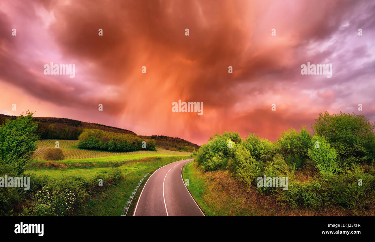 Spectacular rain clouds over a road and green landscape at sunset, with warm red light and dramatic sky Stock Photo