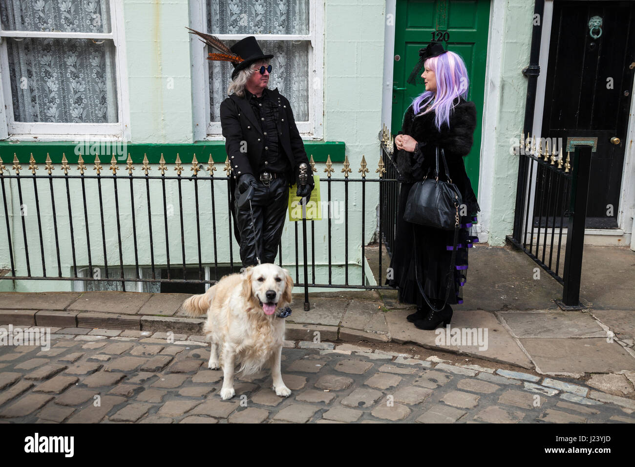 A male and female Goth chat in the street at Whitby ,North Yorkshire at the Goth celebrations accompanied by a Golden Retriever dog on a lead Stock Photo