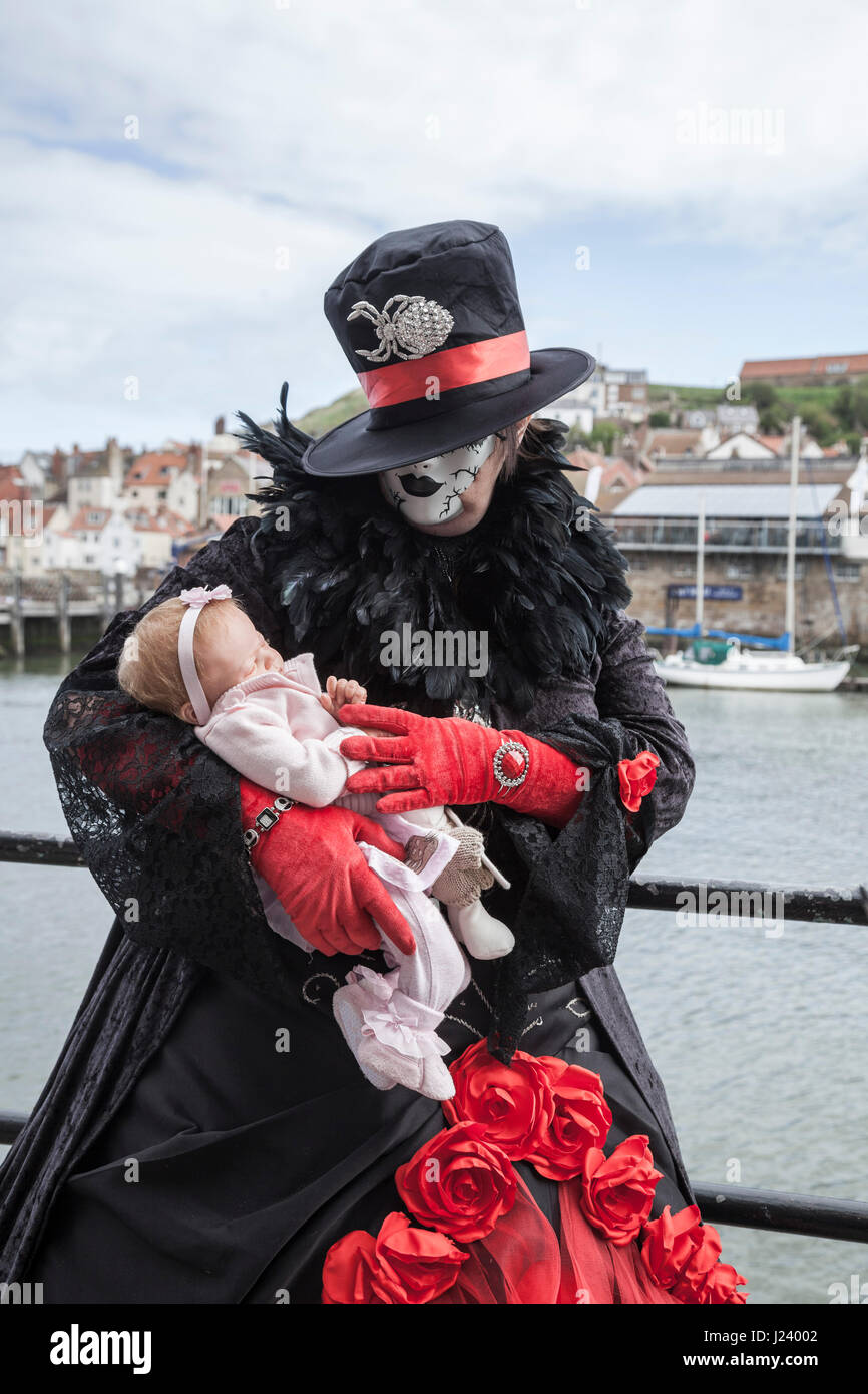 A female Goth stood cradling a lifelike baby doll at Whitby seafront at the biannual Goth Weekend celebrations Stock Photo