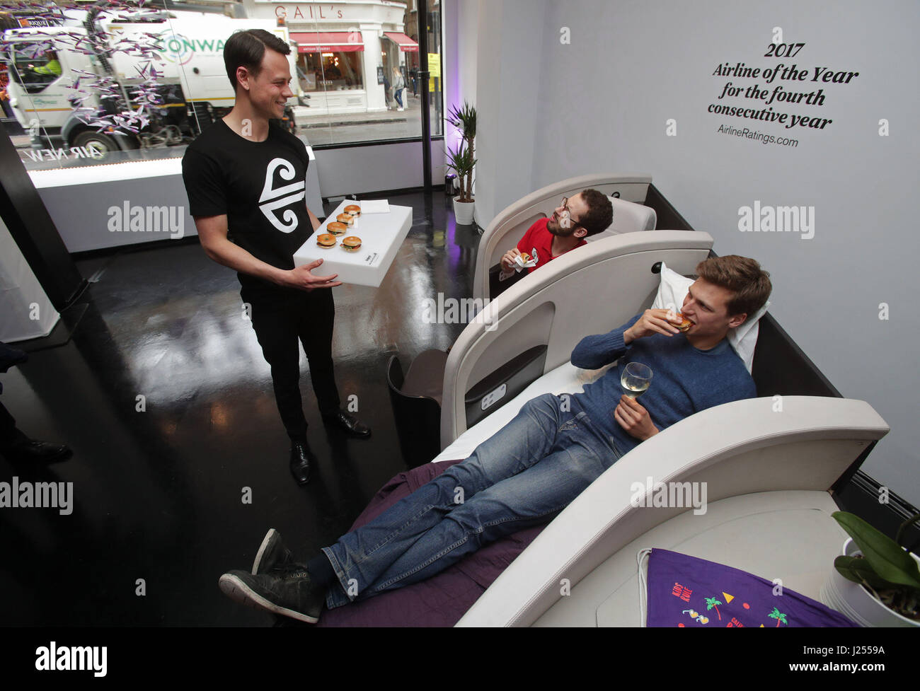 Members of the public Johann Bigler (right) and Fabio Battiato are served snacks by Lachlan McDonald as they sample Business Premier lie-flat seats in a mock-up aircraft cabin, during the launch of Air New Zealands pop-up event 'This is How We Fly', which showcases the airlines on-board facilities, in Soho, London. Stock Photo
