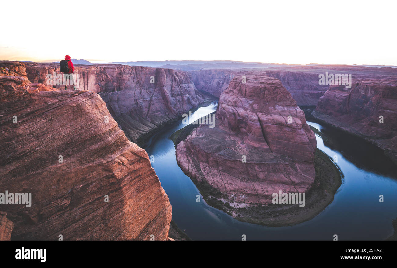 A male hiker is standing on steep cliffs enjoying the beautiful view of Colorado river flowing at famous Horseshoe Bend at sunset, Page, Arizona, USA Stock Photo