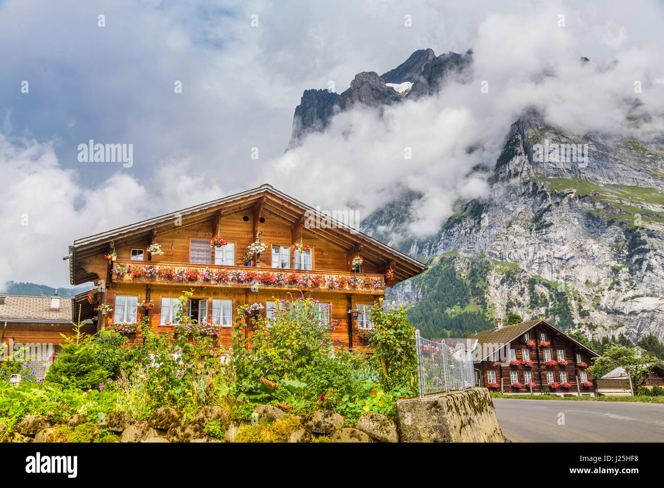 Traditional farmhouses in idyllic mountain scenery with high mountain peaks covered in clouds and fog on a beautiful sunny day in summer, Grindelwald Stock Photo