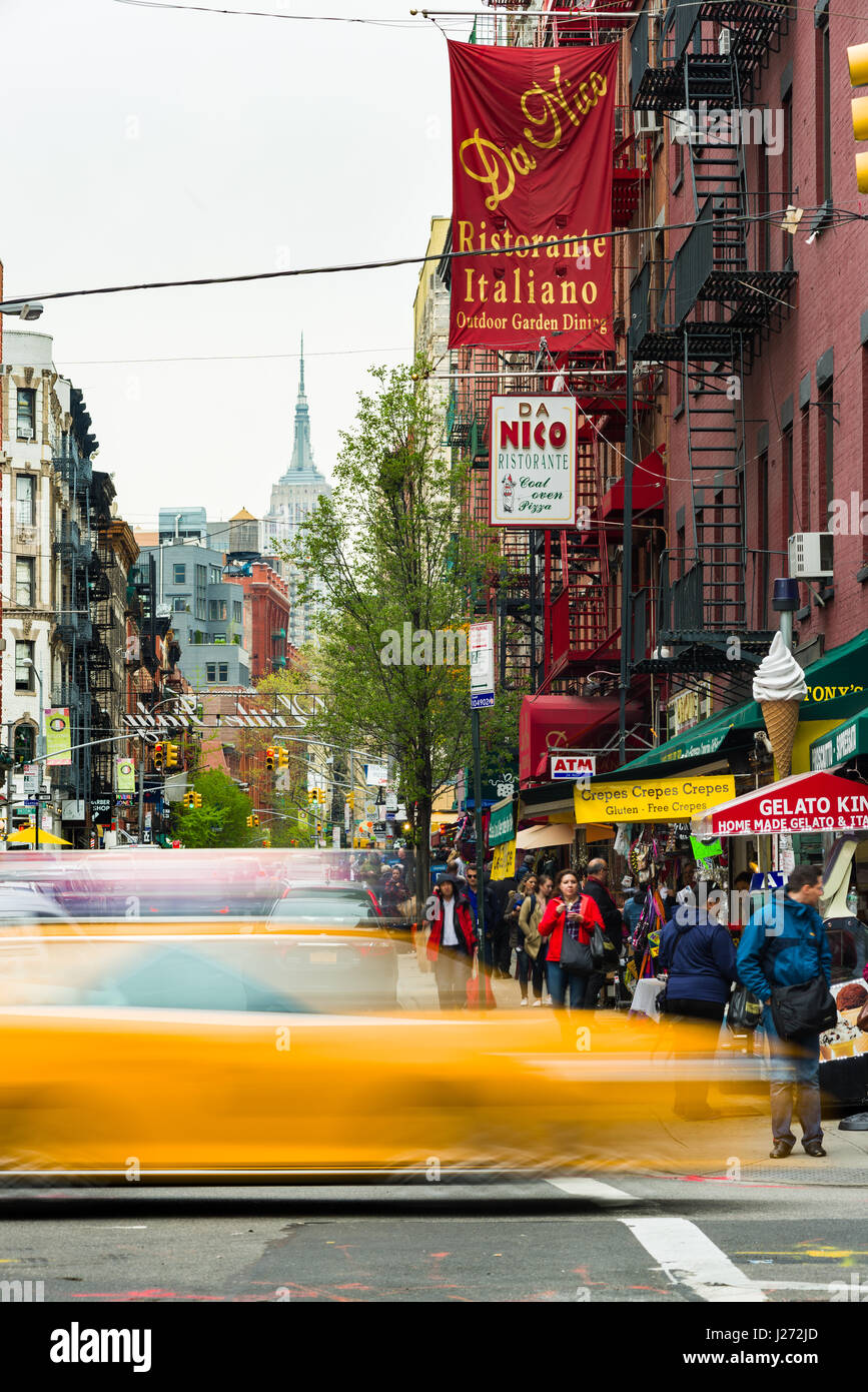 A yellow taxi cab drives past a busy street with restaurants and apartments with the Empire State Building in the background, Little Italy, New York Stock Photo