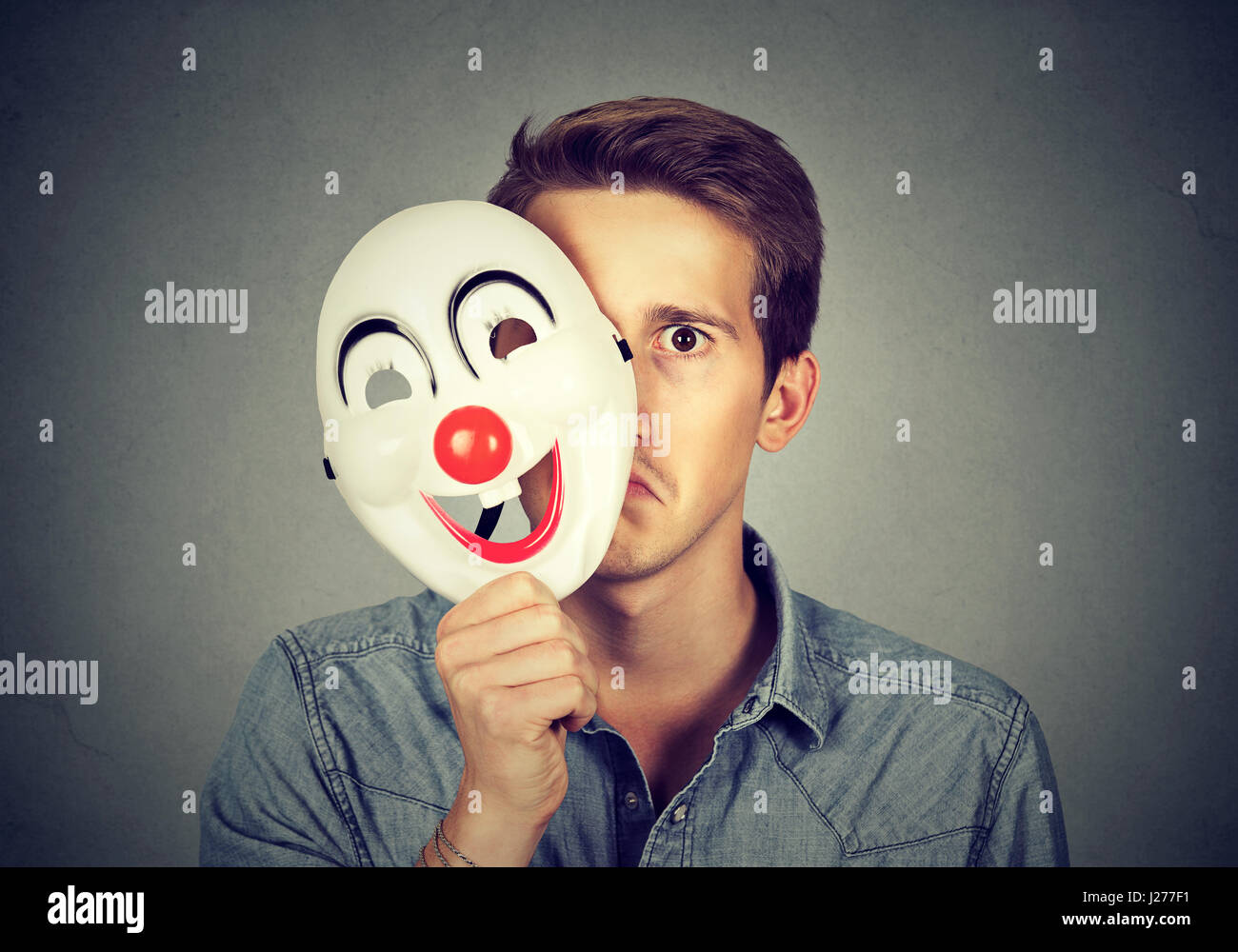 Young sad man hiding behind happy clown mask isolated on gray wall background. Human emotions Stock Photo