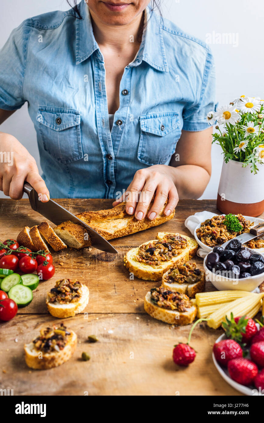 A woman slicing French baguette photographed from front view. Olive tapenade on bread slices, olives, pickled baby corns, strawberries, tomatoes, cucu Stock Photo