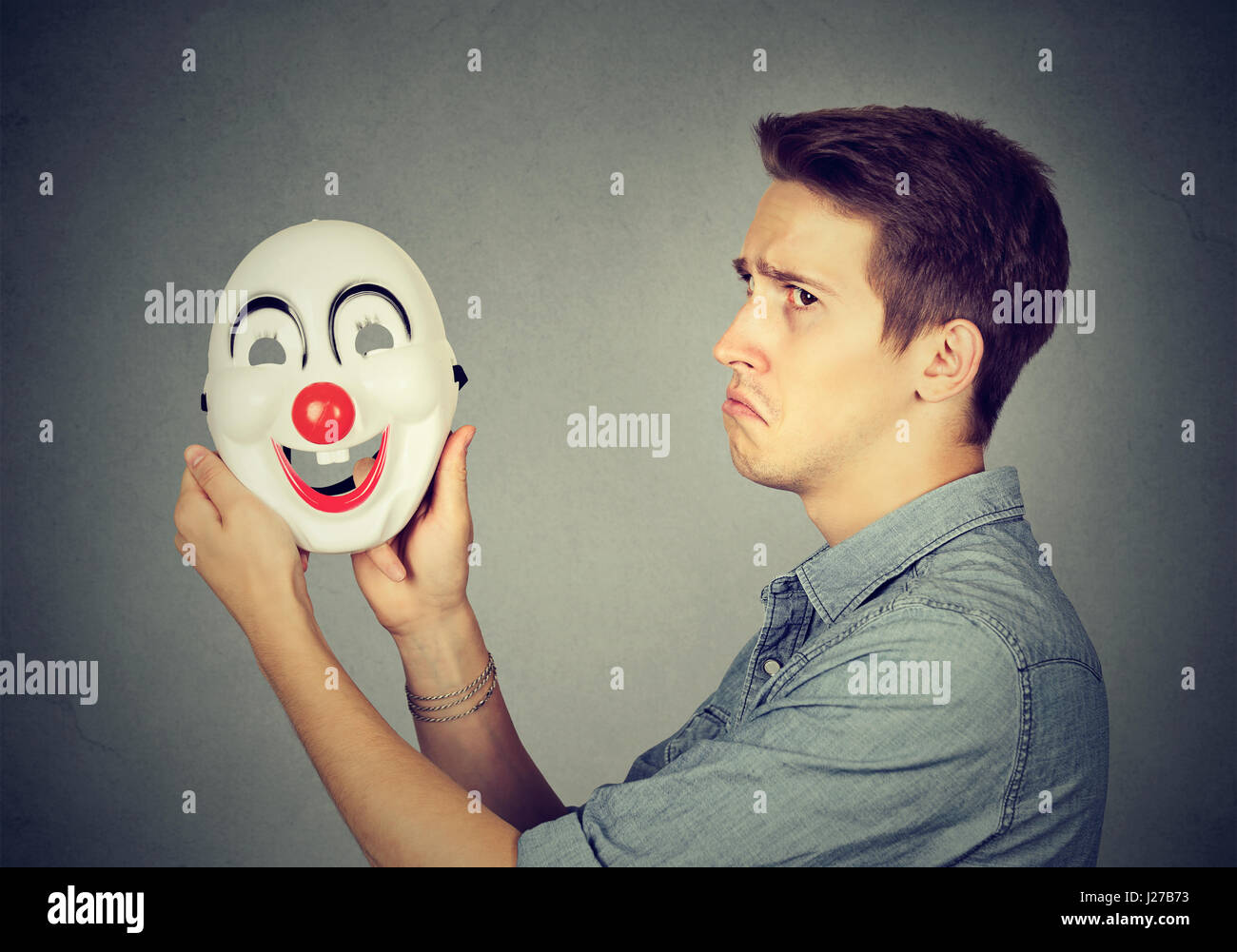 Young sad man with happy clown mask isolated on gray wall background. Human emotions. Split