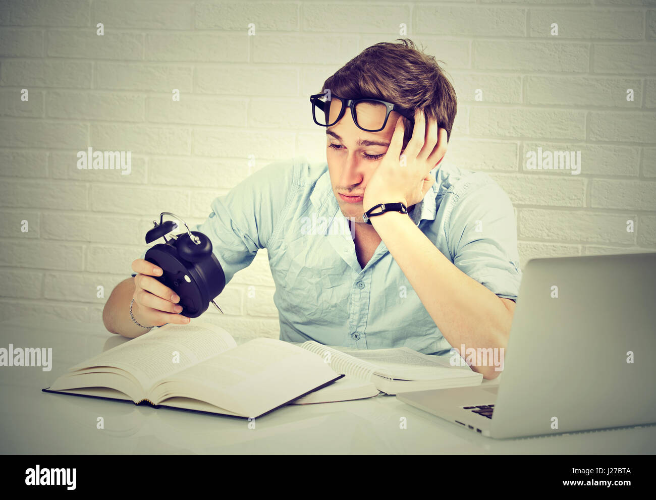 Long studying hours sleep deprivation concept. Tired sleepy young man sitting at desk with book in front of laptop computer looking at alarm clock Stock Photo