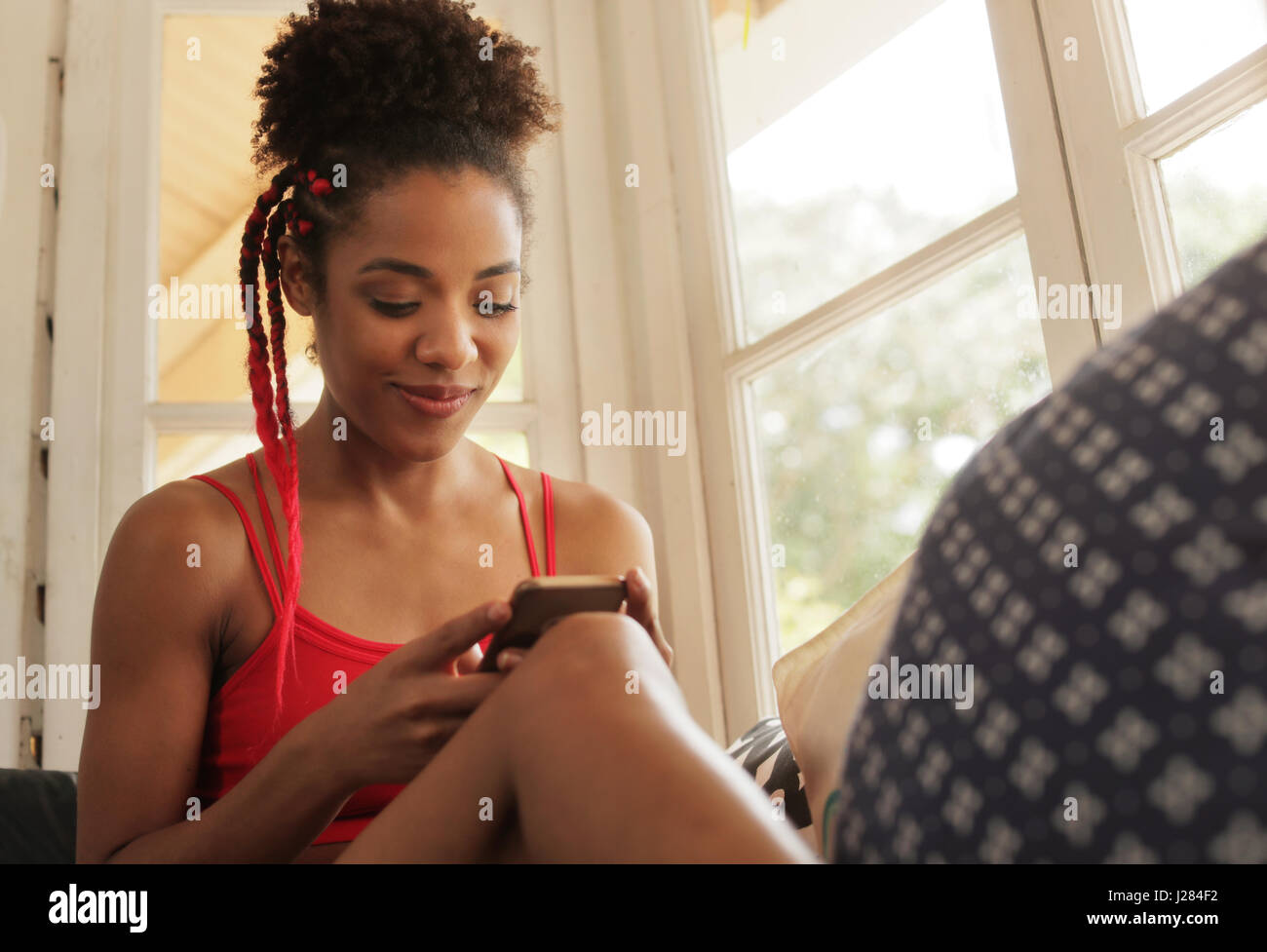 Black girl lying on couch and using smartphone, young african american woman relaxing with mobile phone. Happy latina sitting on sofa, smiling and tex Stock Photo