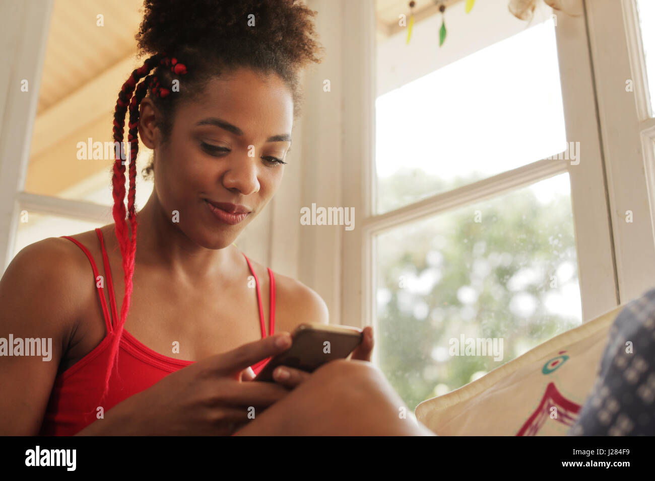 Black girl lying on couch and using smartphone, young african american woman relaxing with mobile phone. Happy latina sitting on sofa, smiling and tex Stock Photo