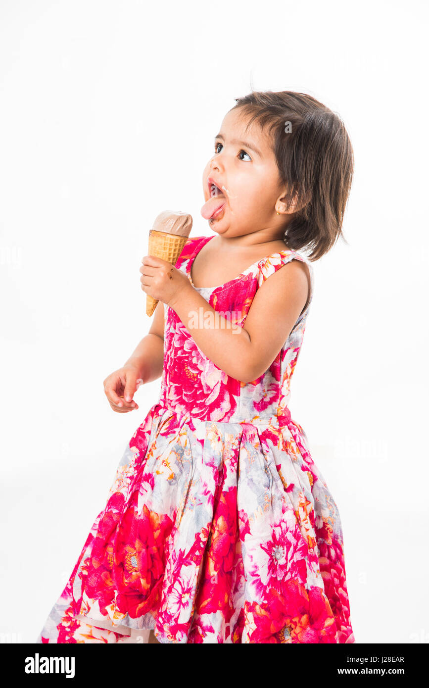 indian adorable infant or girl child licking or eating chocolate ice cream in cone cone and showing happiness, isolated over white background Stock Photo