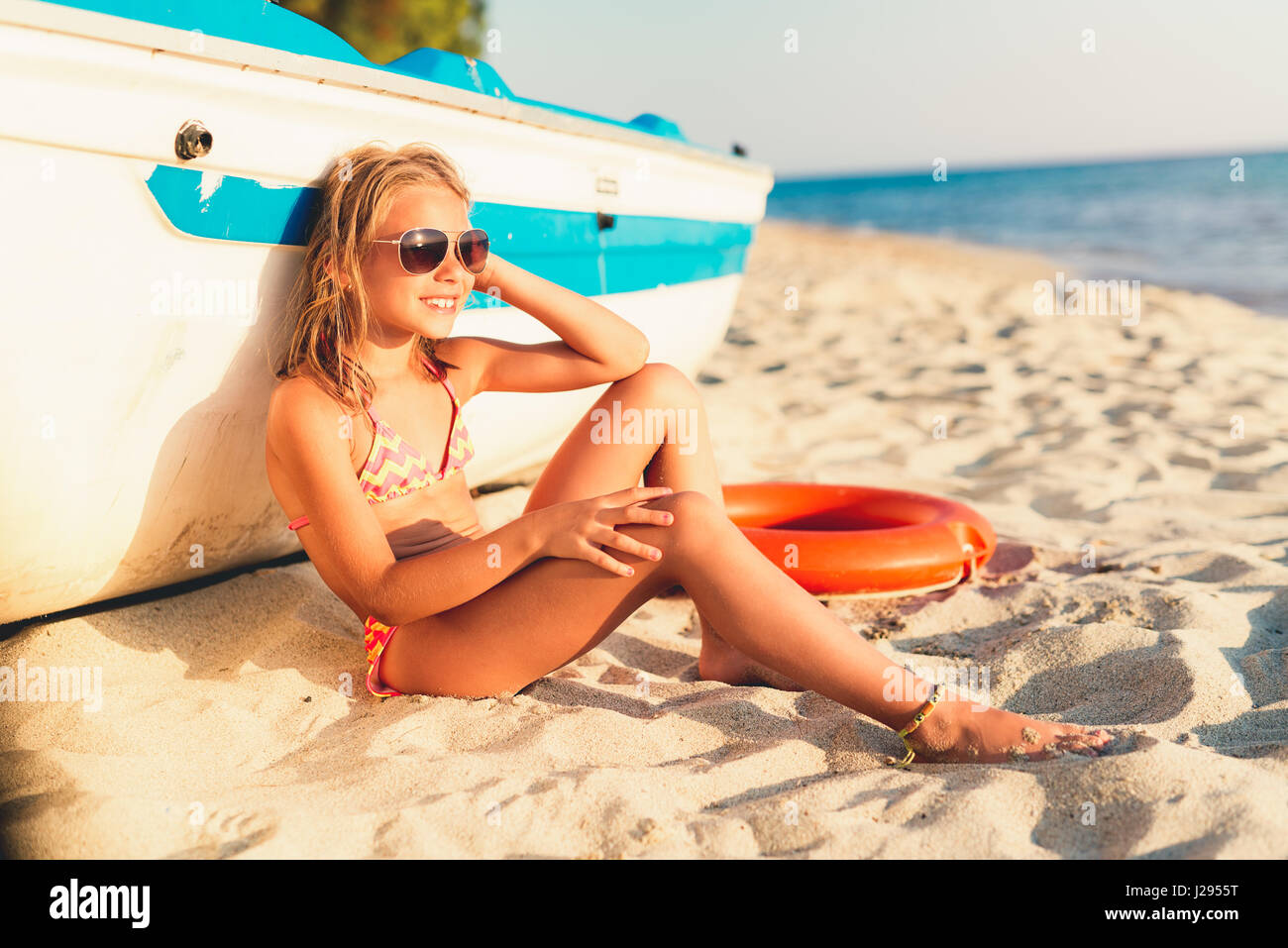 Beautiful little girl with sunglasses relaxing on the beach. She is sitting and sunbathing next to boat. Stock Photo