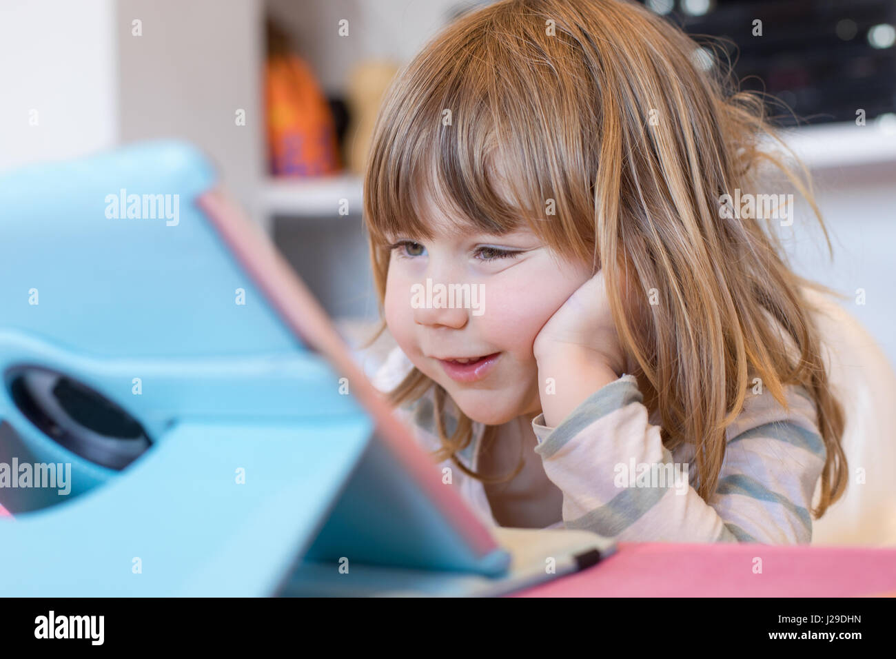smiling face expression of three years old blonde child sitting indoor, with head over hand watching digital tablet on table Stock Photo
