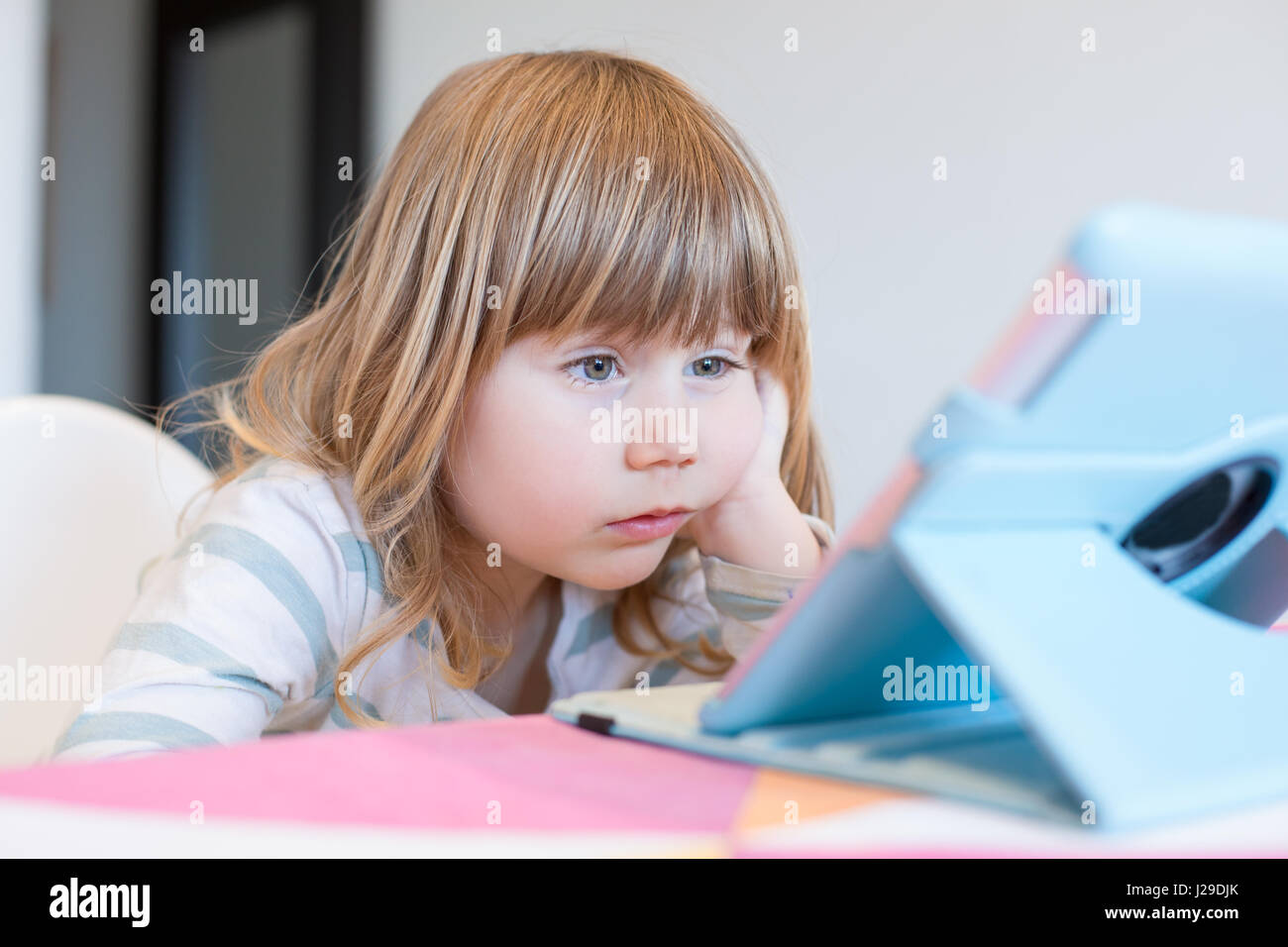 surprise face expression of three years old blonde child sitting indoor, with head over hand watching digital tablet on table Stock Photo