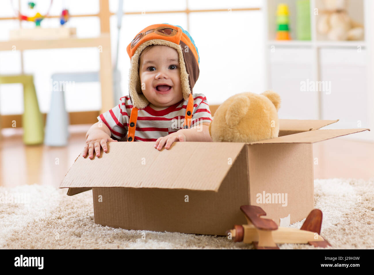 Smiling pilot aviator baby boy with teddy bear toy plays in cardboard box Stock Photo