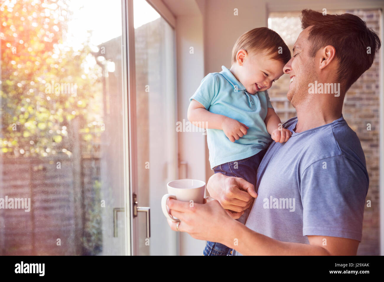 Father holds toddler son while drinking coffee at home, by the window Stock Photo