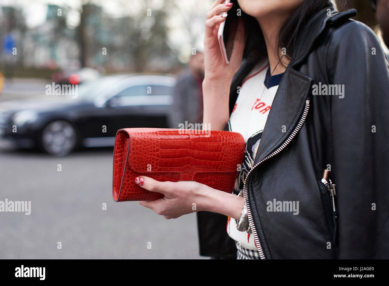 LONDON - FEBRUARY, 2017: Mid section close up of woman wearing a black leather bike jacket, holding a red Hermes purse and using smartphone in the street during London Fashion Week, horizontal Stock Photo