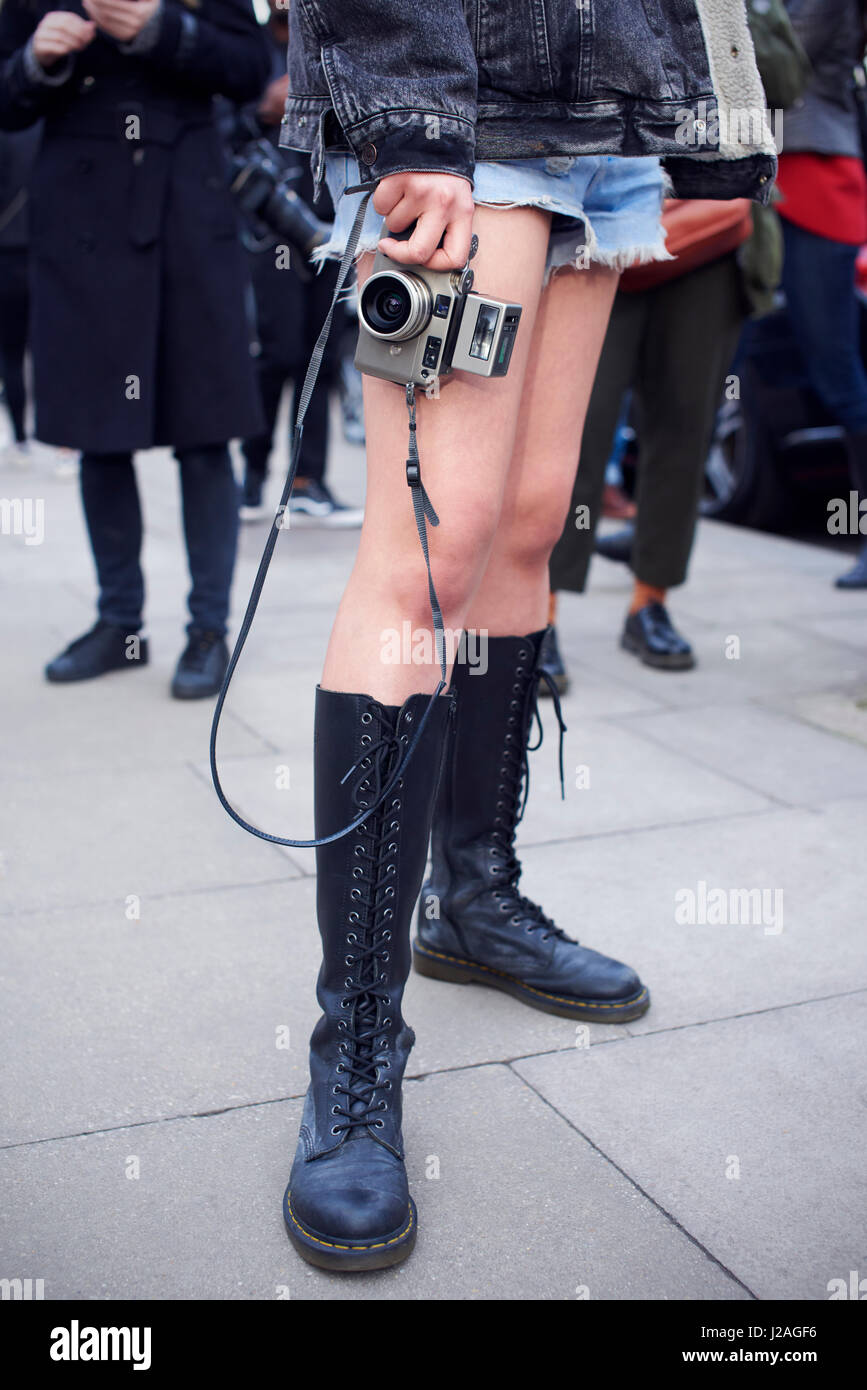 LONDON - FEBRUARY, 2017: Low section of woman wearing tall Dr Martens boots and denim shorts holding a camera in the street during London Fashion Week, vertical, front view Stock Photo