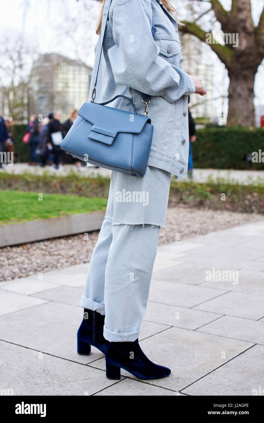 LONDON - FEBRUARY, 2017: Low section of woman wearing grey denim jacket and jeans with a blue leather shoulder bag standing in the street during London Fashion Week, vertical, side view Stock Photo